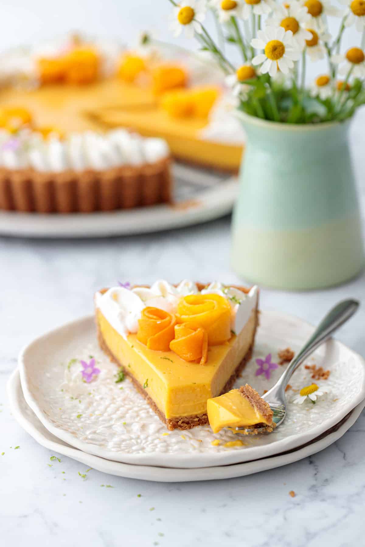 Slice of Mango Lime Tart on a plate with a forkful on the side to show the creamy texture, with vase of flowers and rest of the tart in the background.