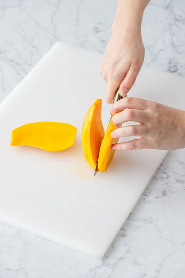 On a white cutting board, slicing the cheeks off of a peeled mango.