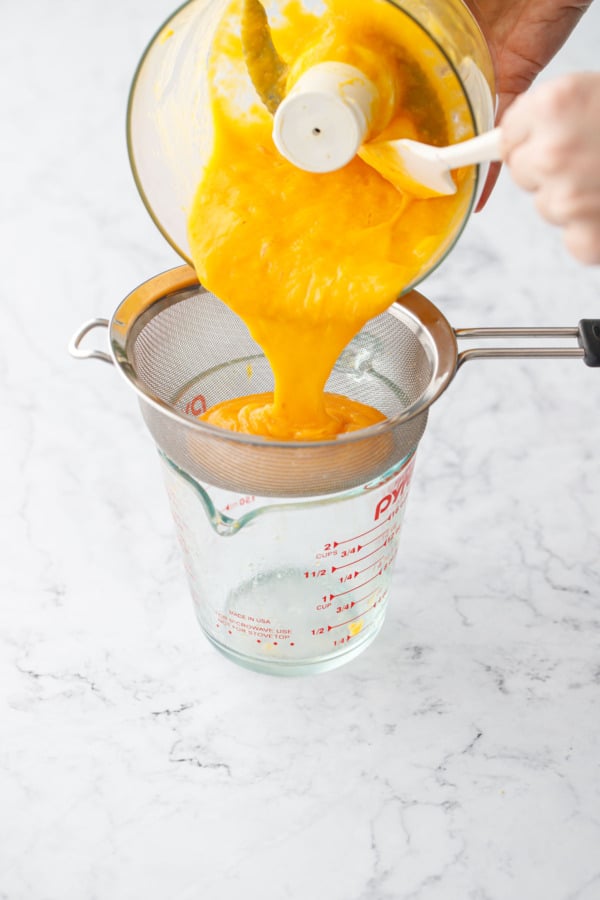 Pouring pureed mango from a food processor into a mesh sieve set over a glass measuring cup.