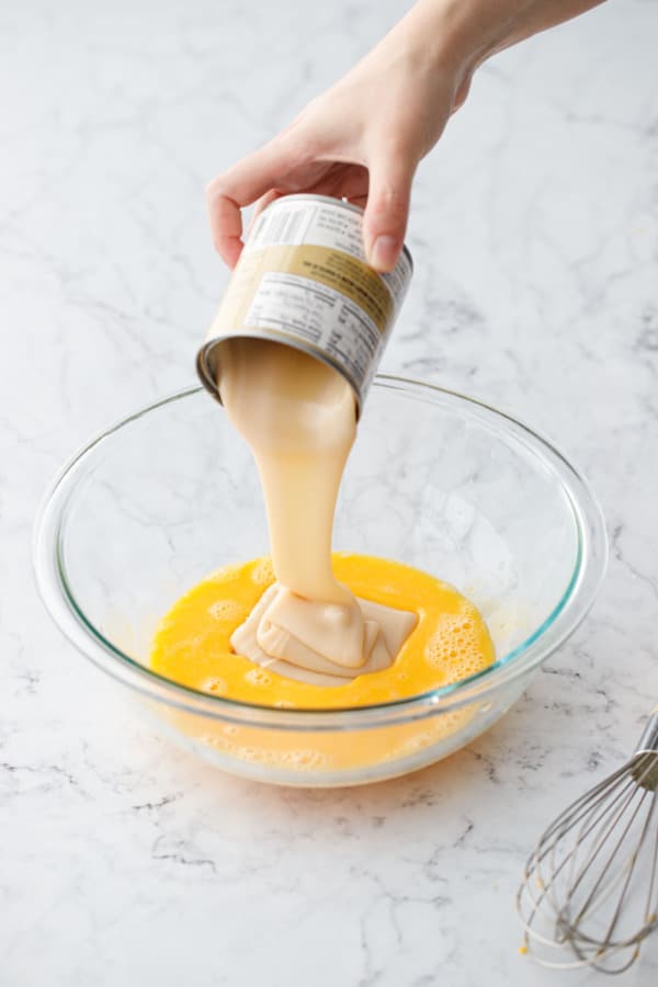 Pouring sweetened condensed milk into a mixing bowl with eggs and lime juice.