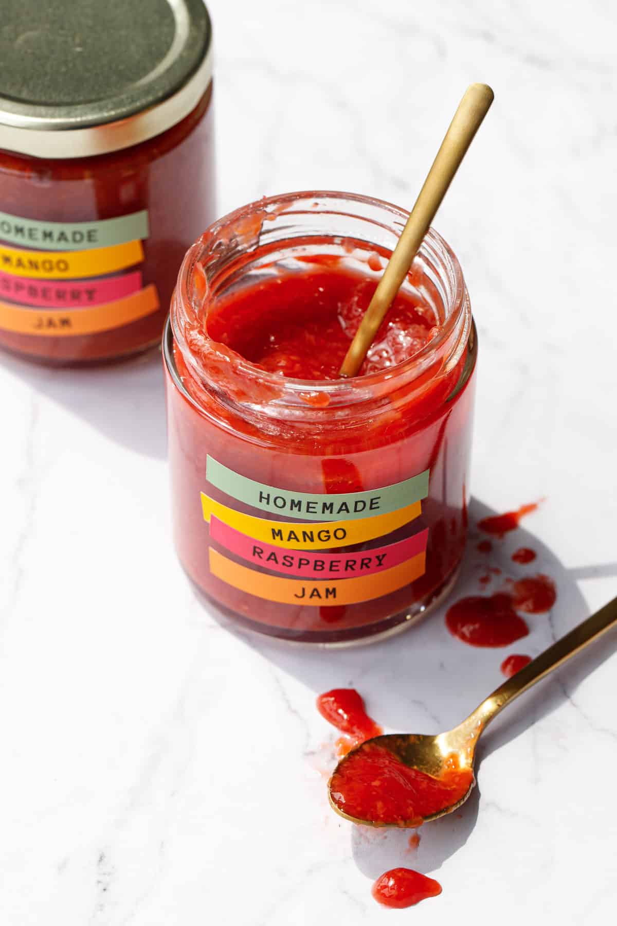Two jars of Mango Raspberry Jam, gold spoon with spilled jam smeared on the marble background.