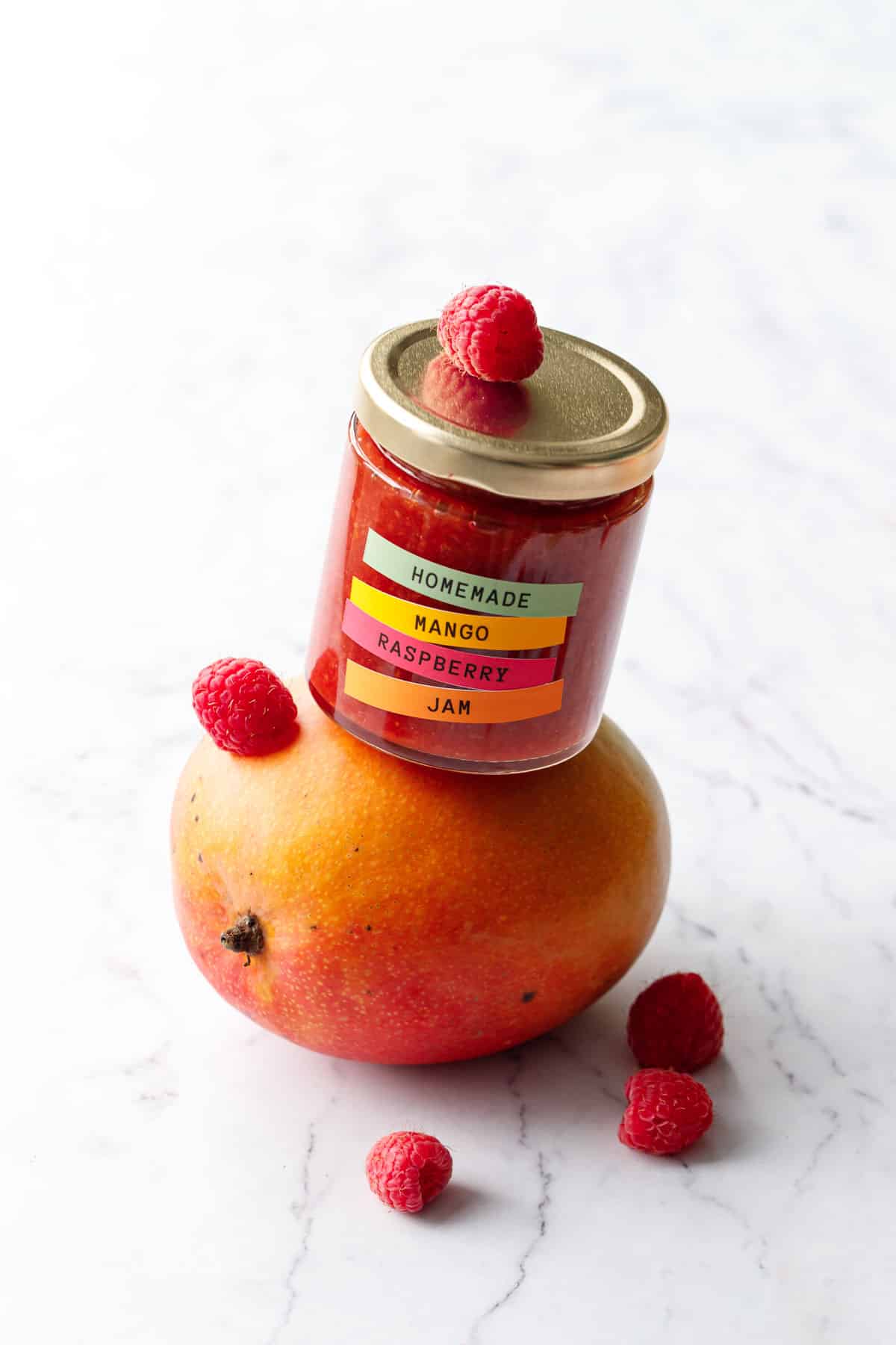 Jar of Mango Raspberry Jam balancing on a mango with a few raspberries scattered around.