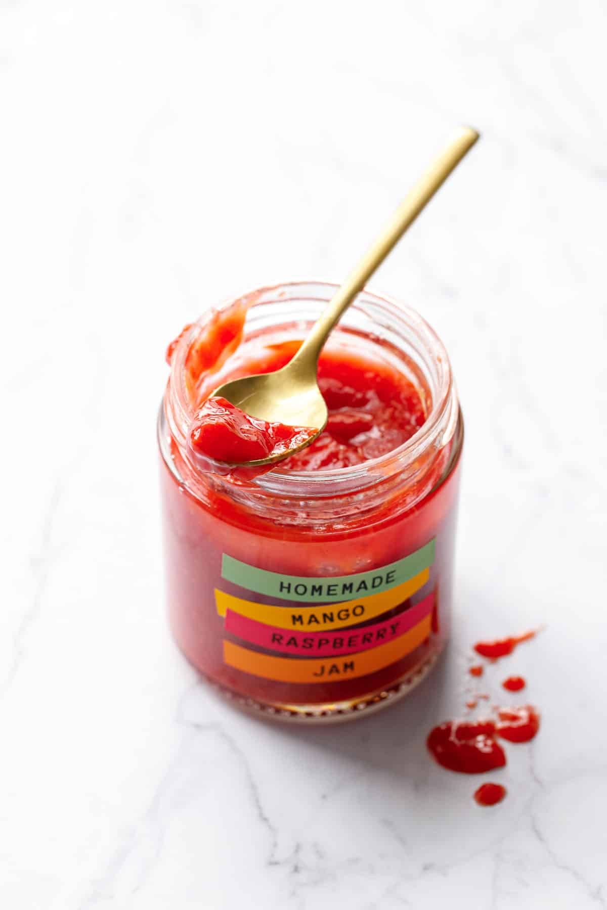 Open jar of Mango Raspberry Jam with a gold spoon propped on top full of jam to show the smooth texture.