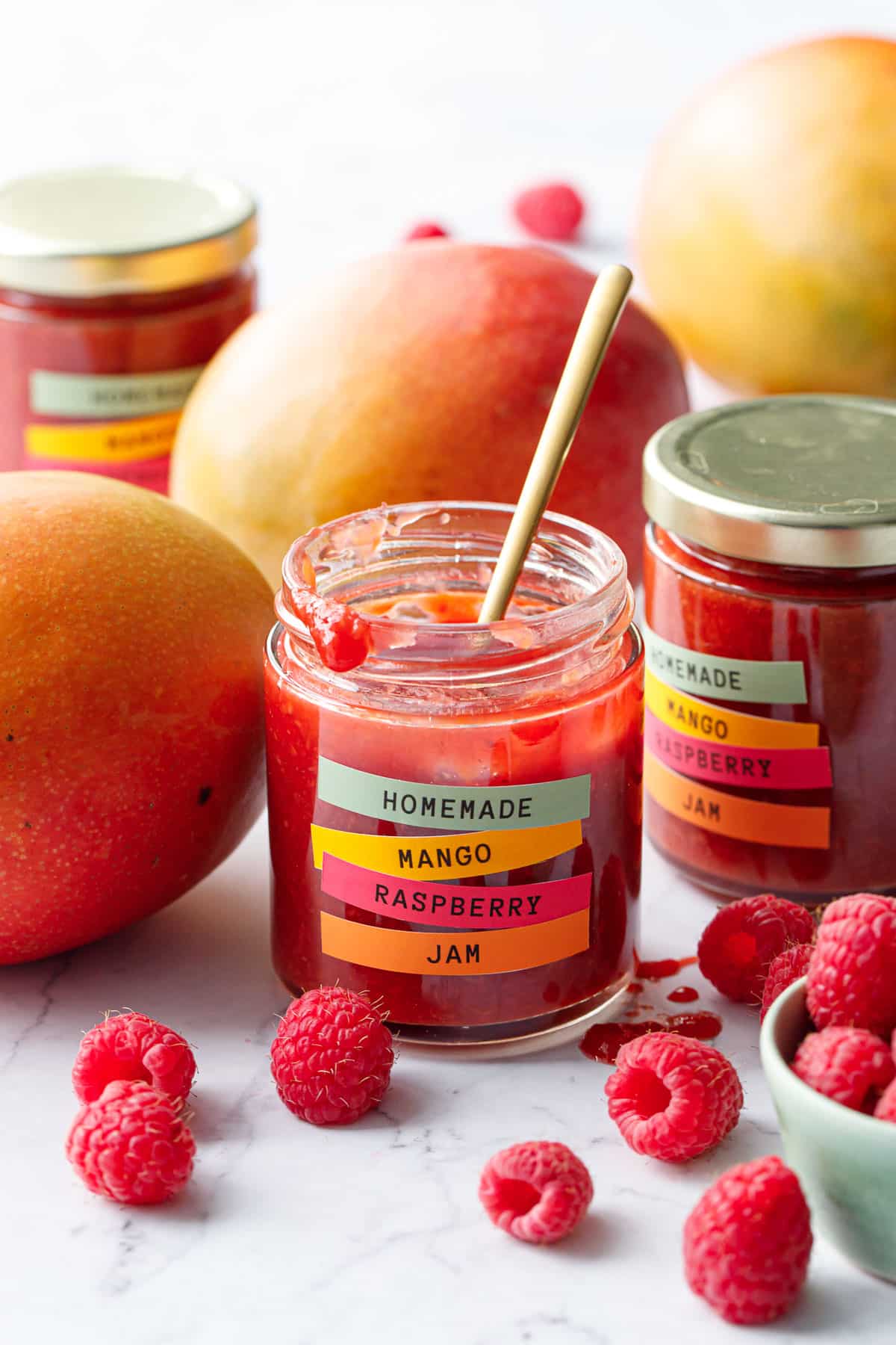 Jars of Mango Raspberry Jam with fresh mangoes and raspberries scattered around, one jar open with a spoon sticking out.