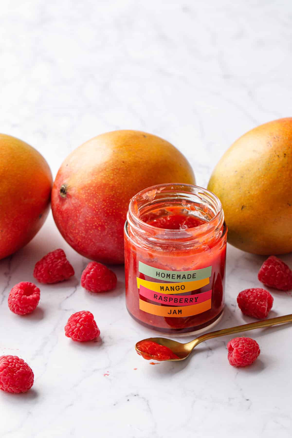 Row of mangoes with scattered raspberries and a jar of Mango Raspberry Jam in front, with a gold spoon filled with jam.