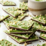 Stack of cut pieces of Matcha White Chocolate Crunch Bark showing the layers, cup with matcha latte and bowl of matcha powder in the background