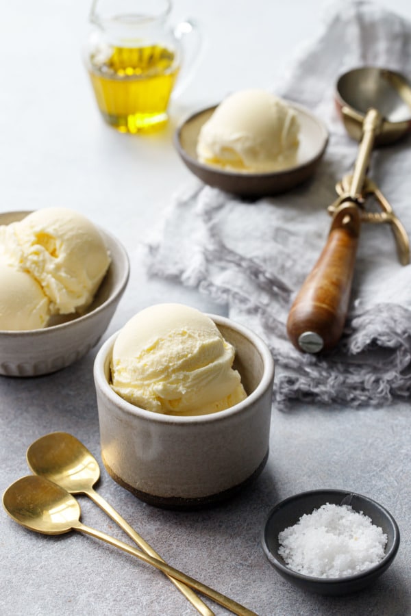 Scoops of olive oil ice cream in gray ceramic dishes with a vintage ice cream scoop and bowl of fleur de sel
