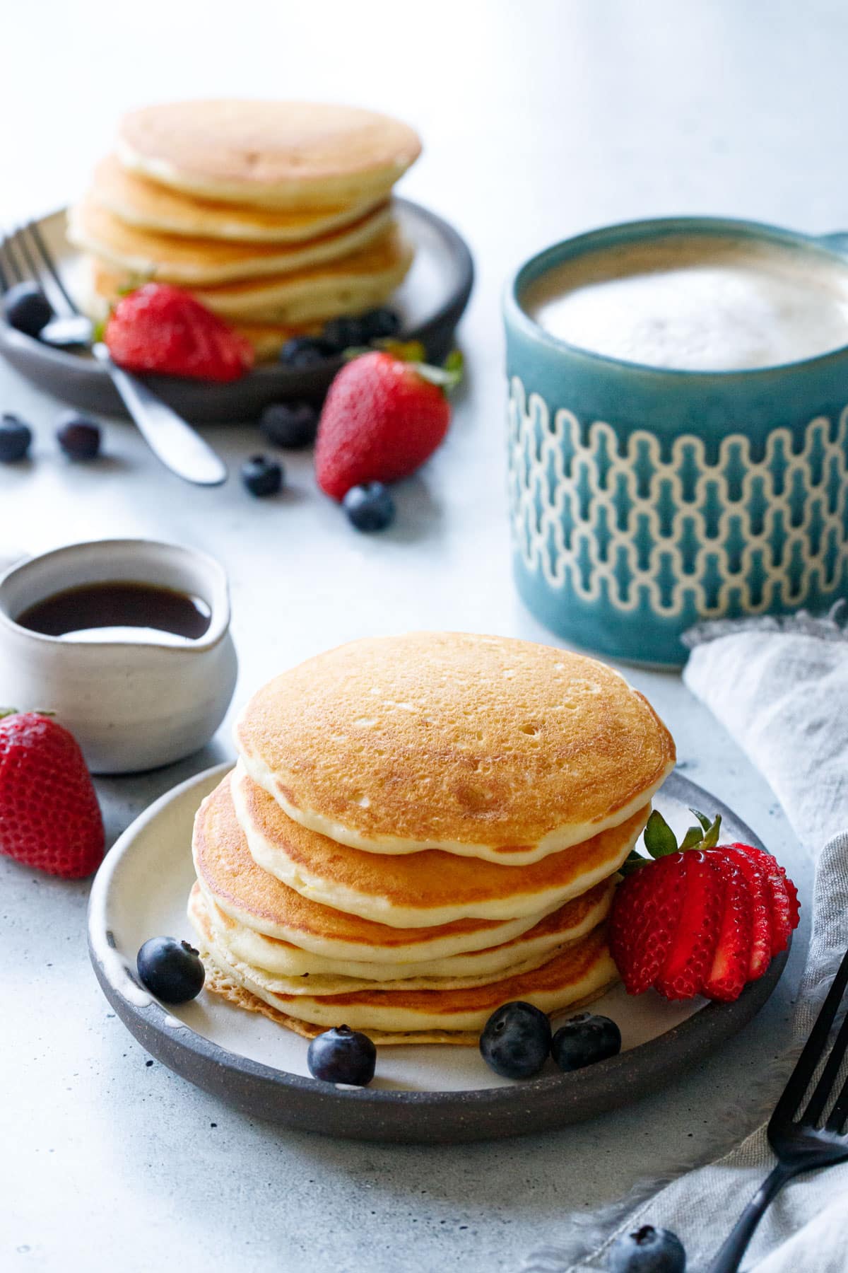 Two stacks of Olive Oil Pancakes on ceramic plates with strawberries and blueberries, mug of coffee on the side.