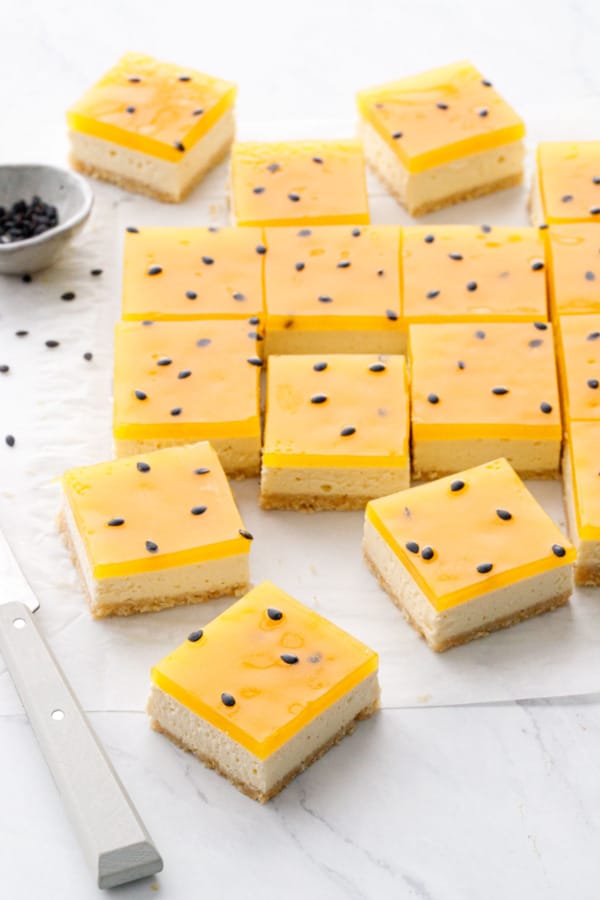 Passionfruit Cheesecake Bars cut into squares, on a piece of white parchment with a gray paring knife and bowl of passionfruit seeds in the background.