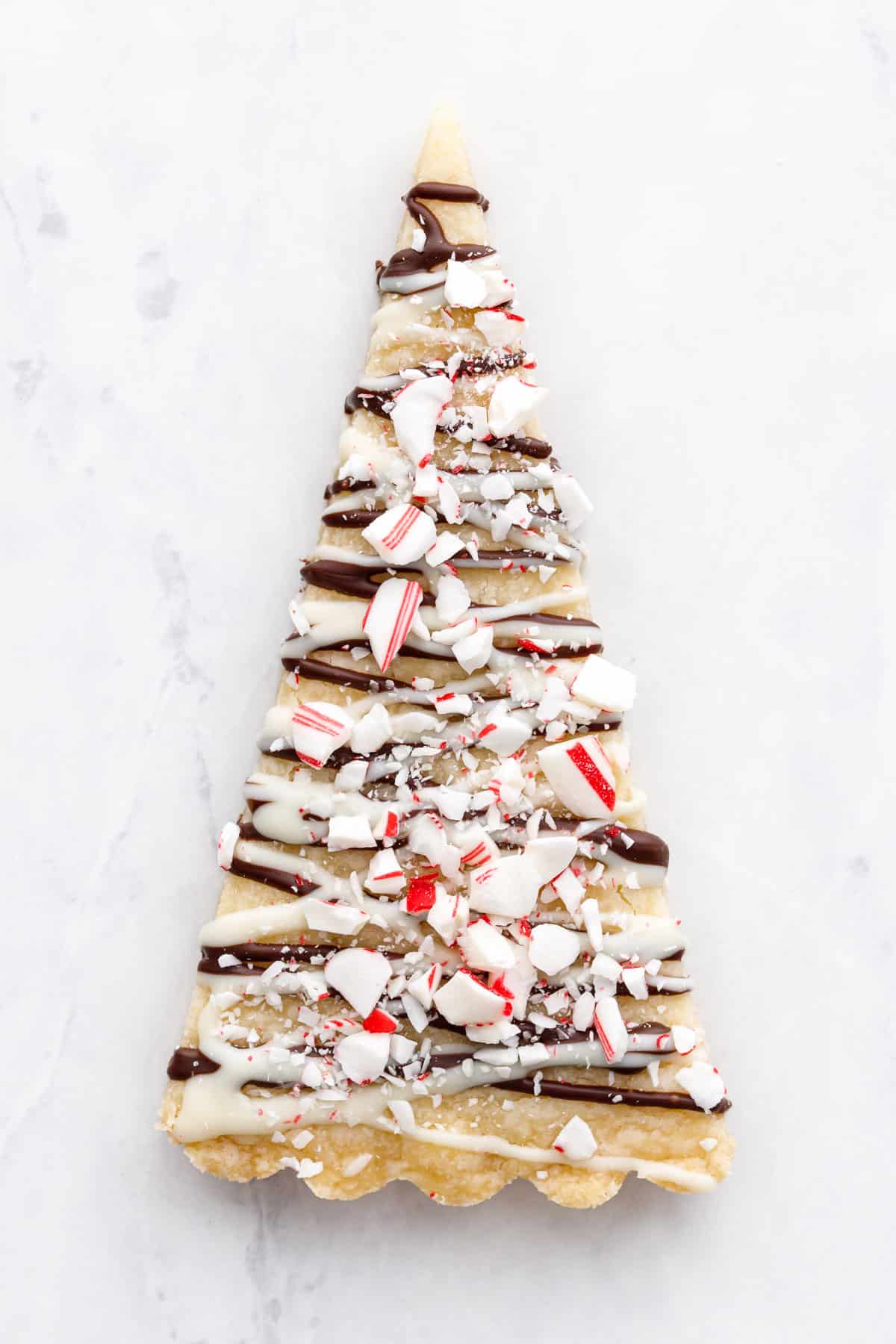 Peppermint Bark Shortbread cut into triangles to look like Christmas trees, decorated with drizzled white and dark chocolate and crushed candy canes.