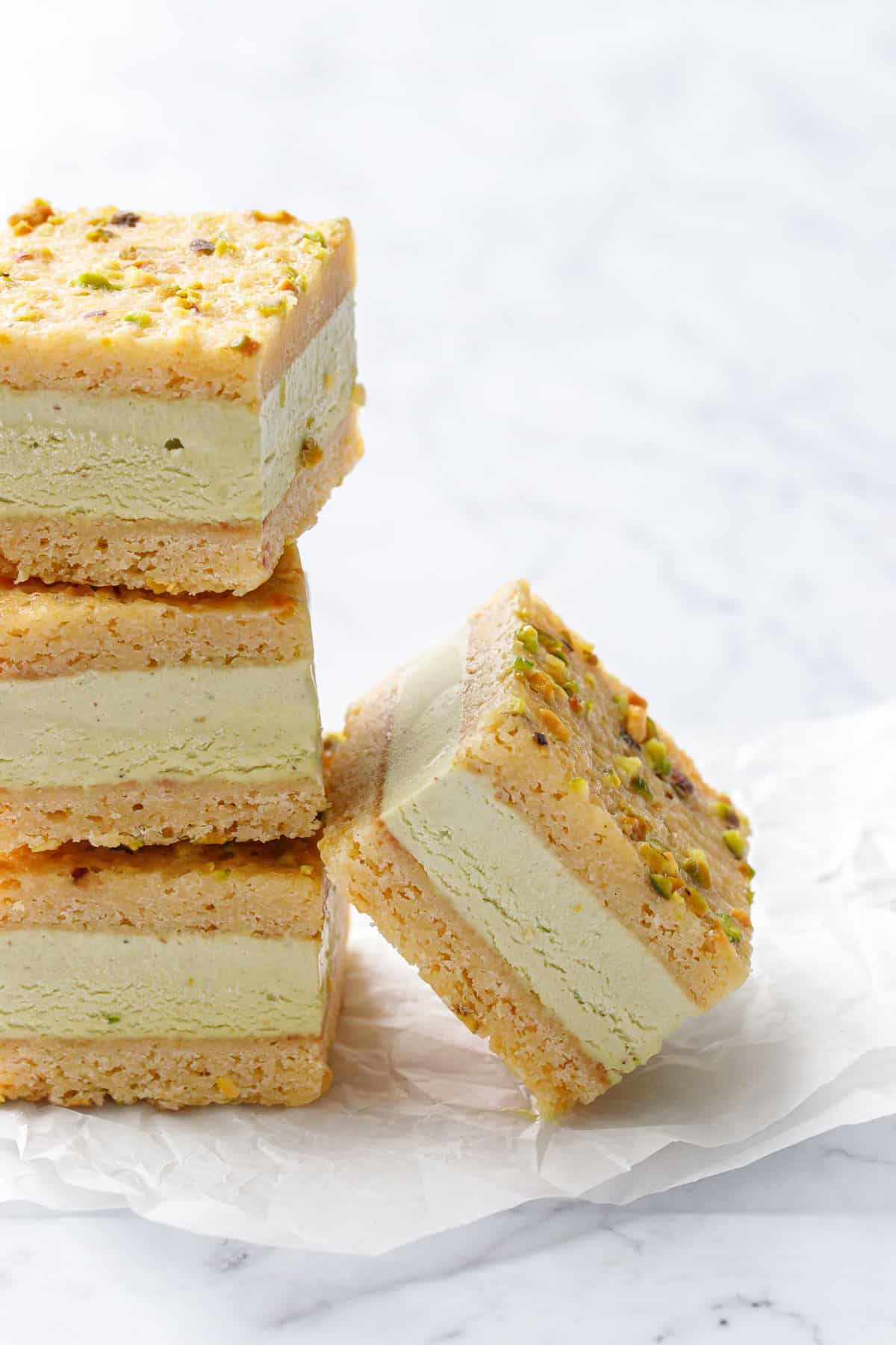 Stack of three Pistachio Blondie Ice Cream Sandwiches with one more square leaning against the side on a white marble background and crinkled square of parchment paper.