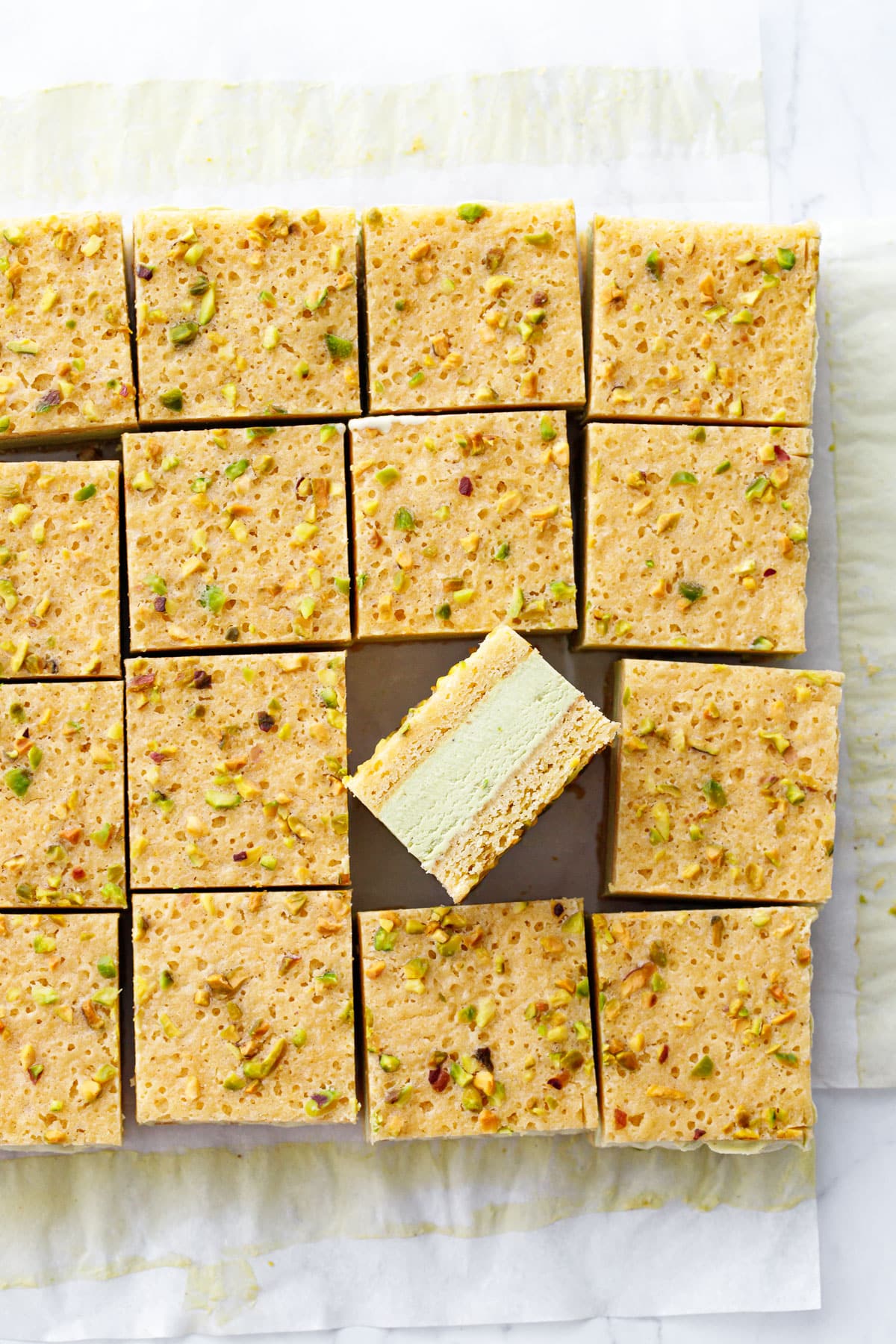 Overhead, cut squares of Pistachio Blondie Ice Cream Sandwiches on parchment, one square propped on its side to show the layers.