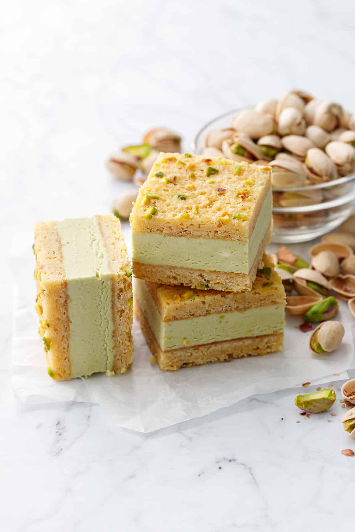 Three squares of Pistachio Blondie Ice Cream Sandwiches stacked on a marble background with bowl of pistachios on the side.
