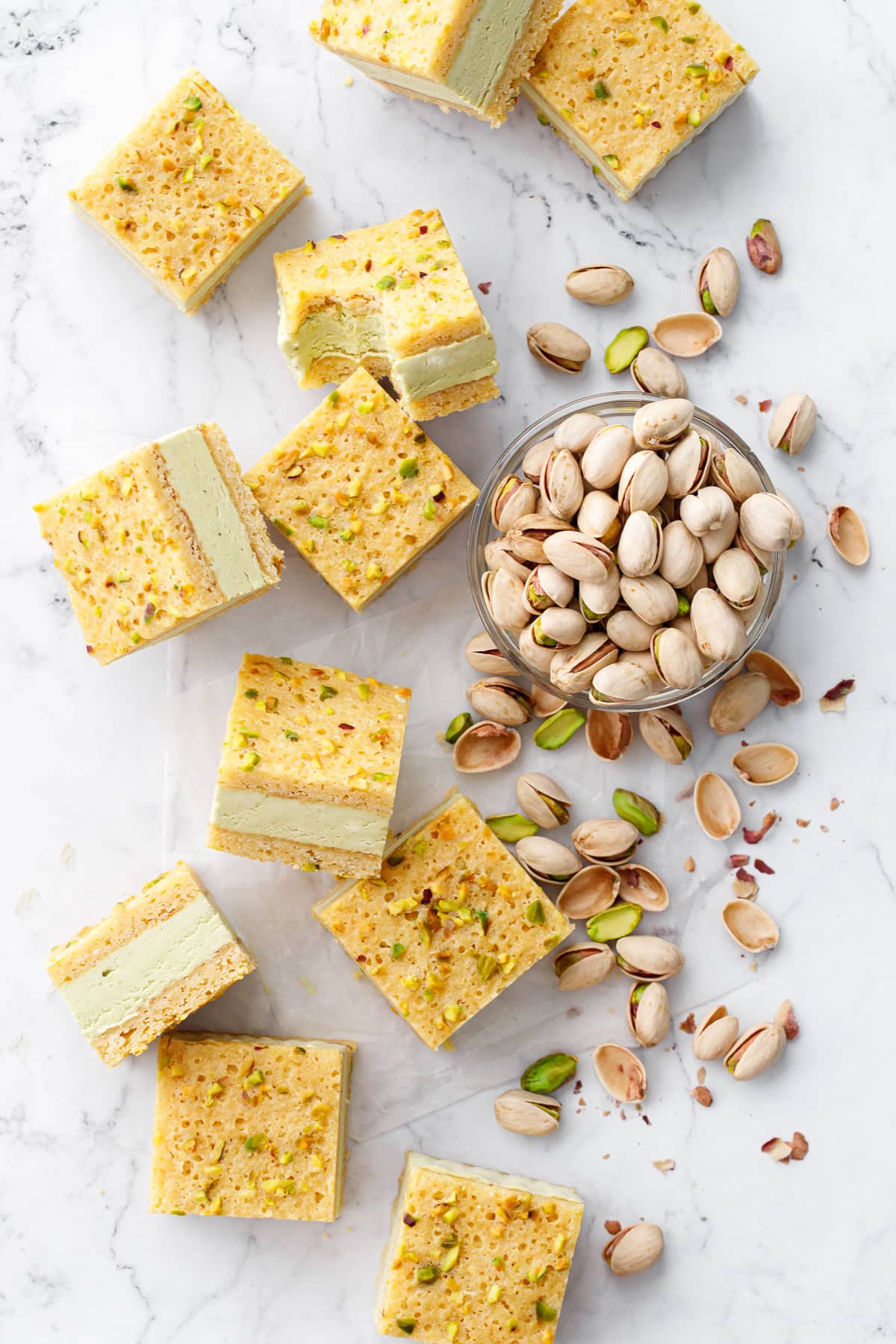 Overhead, messy arrangement of Pistachio Blondie Ice Cream Sandwiches cut into squares, with bowl of pistachios and more pistachios scattered around.