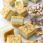 Cut squares of Pistachio Blondie Ice Cream Sandwiches scattered on a marble background with a bowl of pistachios on the side.