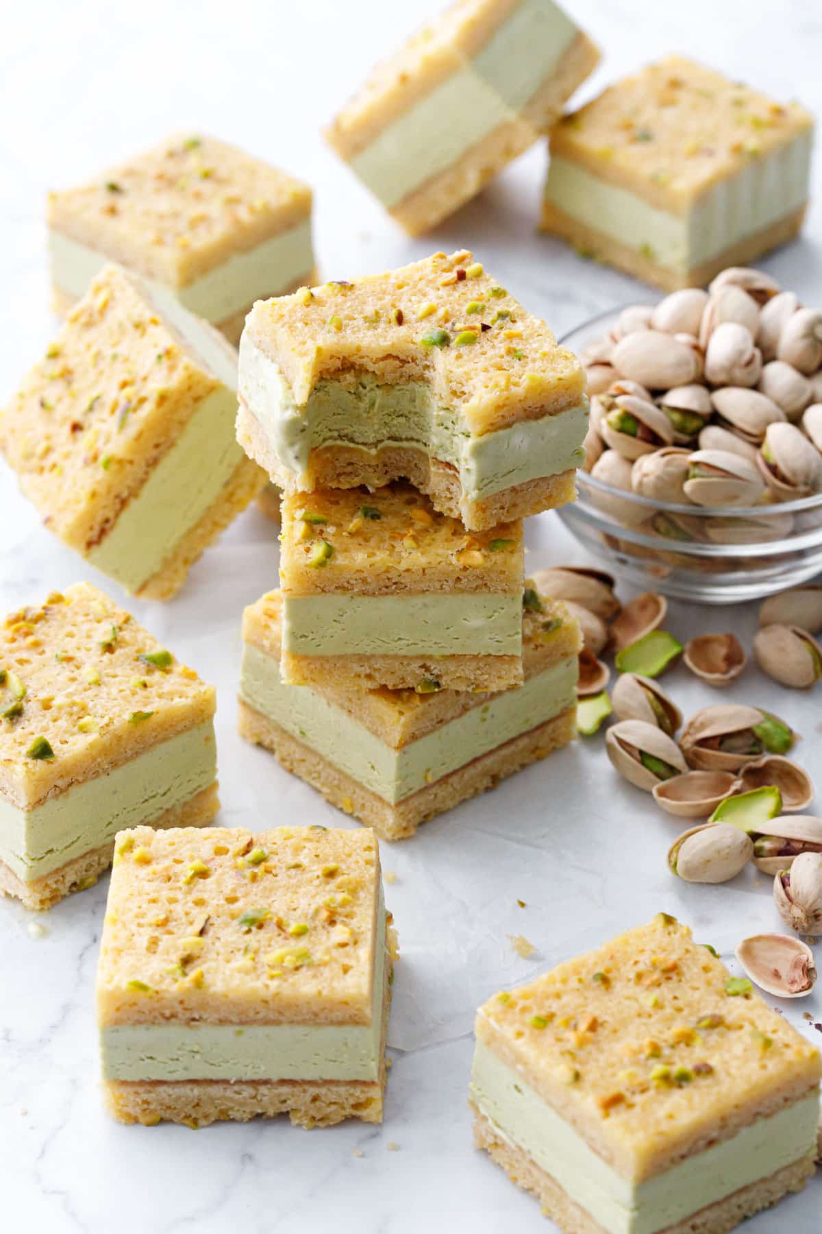 Stack of Pistachio Blondie Ice Cream Sandwiches, one with a bite cut out of it to show the texture, with a bowl of pistachios on the side.