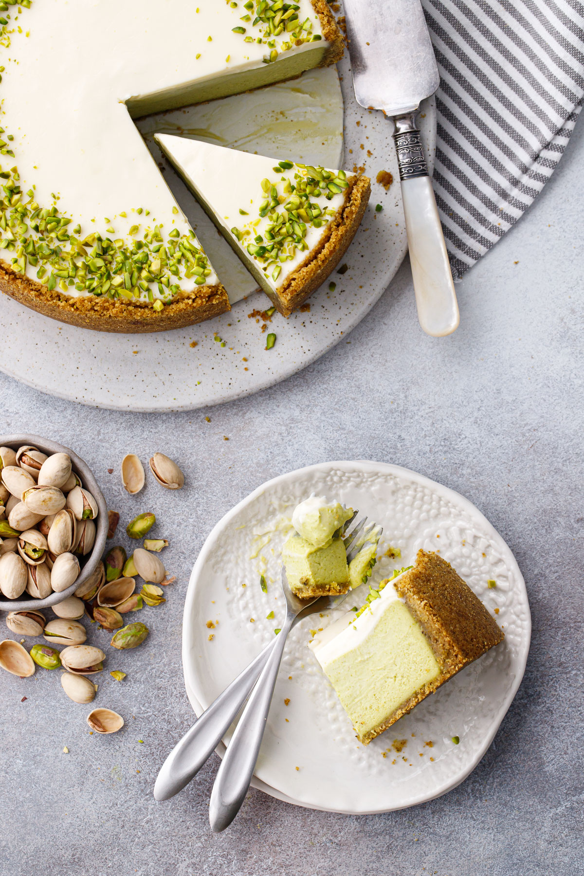 Overhead, slice of Pistachio Sour Cream Cheesecake on its side on a white plate with a forkful on the site, with bowl of messy pistachios and cake plate with the full cheesecake.