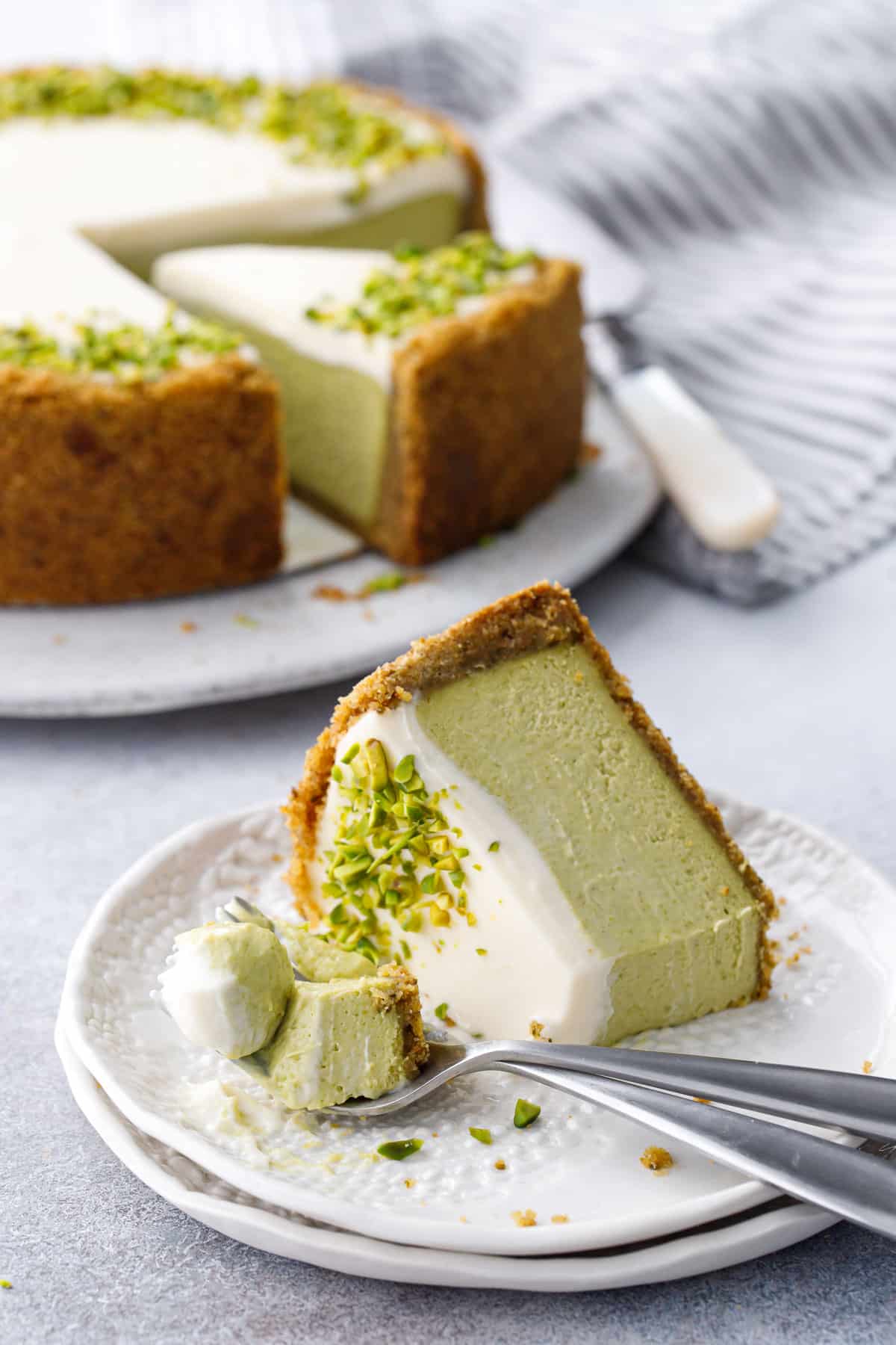 Slice of Pistachio Sour Cream Cheesecake on its side on a white plate, with two forks and a bite of cheesecake, full cheesecake in the background.