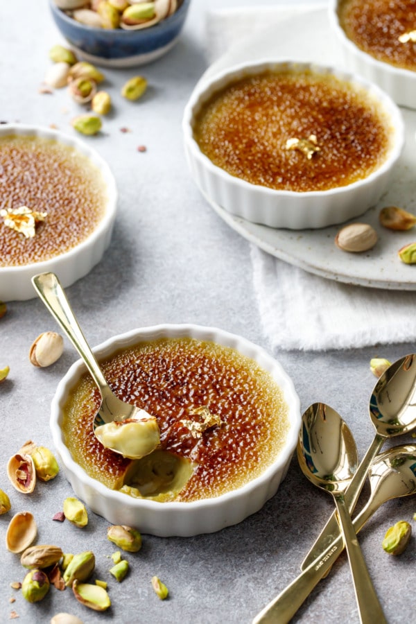 White ramekins with Pistachio Crème Brûlée, topped with a speck of gold leaf, one ramekin with a spoonful taken out to show the creamy texture.
