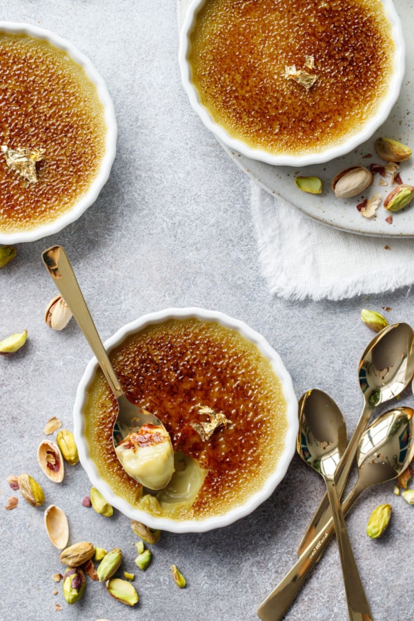 Overhead, three white ramekins filled with Pistachio Crème Brûlée on a gray background with gold spoons and fresh shelled pistachios.