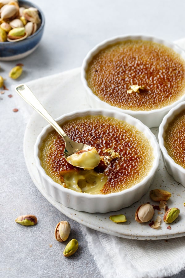 Pistachio Crème Brûlée in fluted ramekins, one with a spoonful out of it to show the creamy texture, with messy pistachios scattered around.