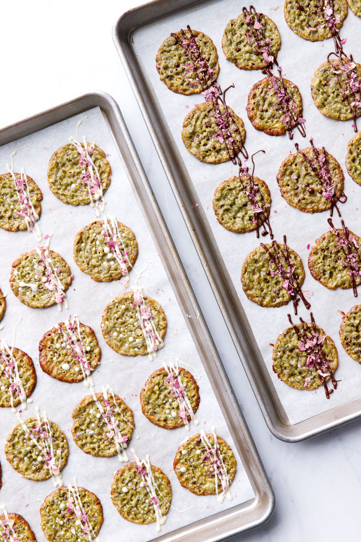 Two sheet pans of Pistachio Florentine Cookies on the diagonal, one with dark-chocolate drizzle and one with white chocolate.