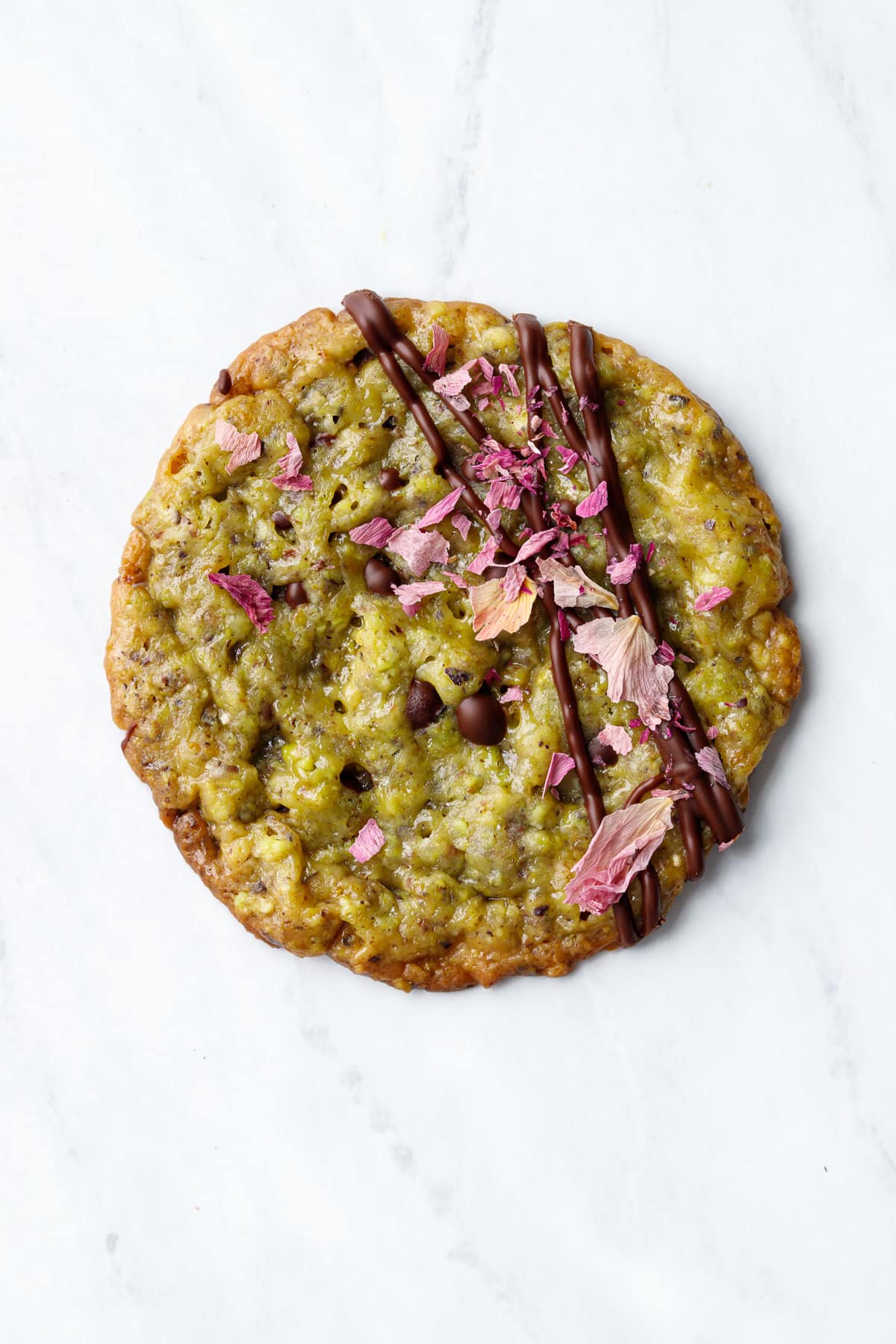 Closeup, single Pistachio Florentine Cookie on a marble background, drizzled with off-center stripes of dark chocolate and sprinkled with dried rose petals.