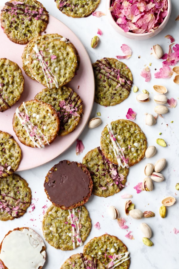 Overhead, messy scene with Pistachio Florentine Cookies on a pink plate, some upside down to show the chocolate and white-chocolate coated bottoms, with scattered pistachios and rose petals.