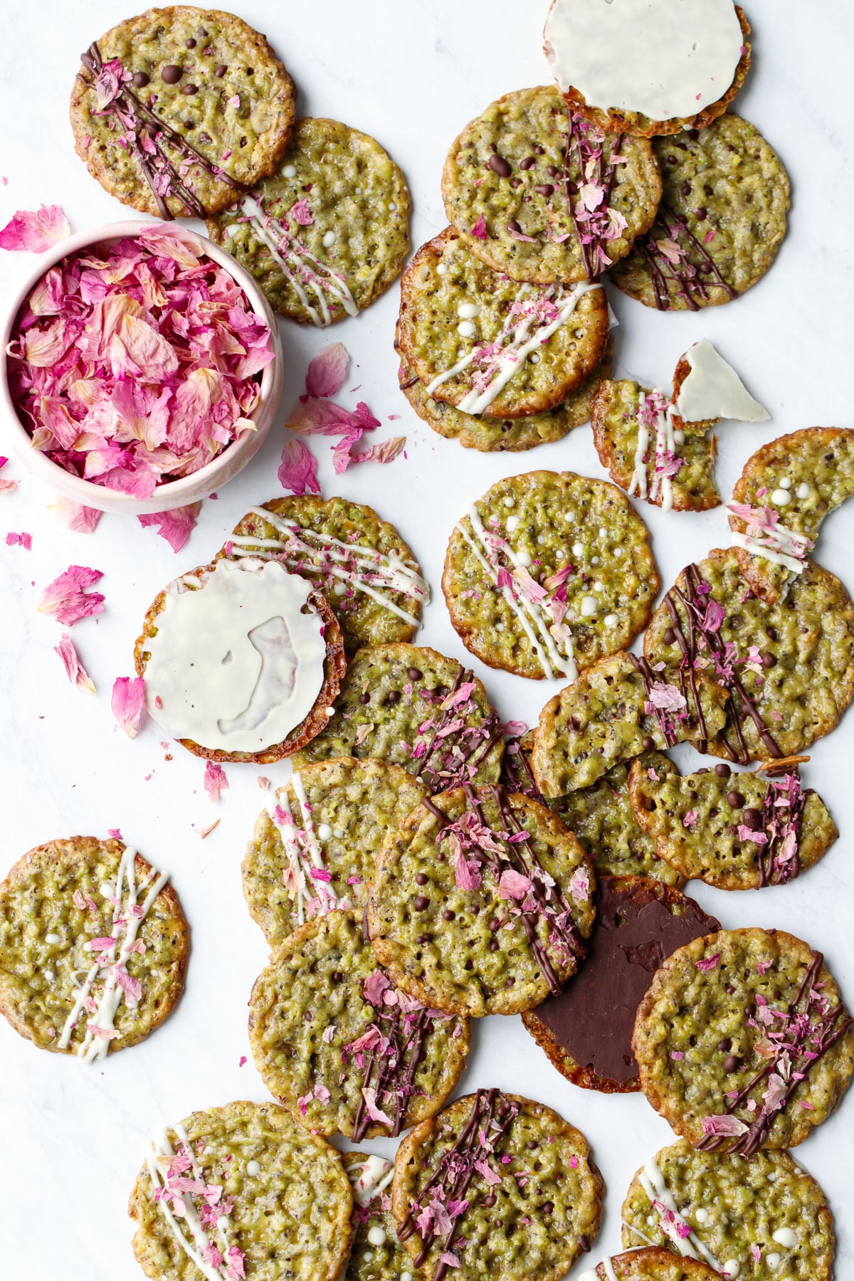 Overhead, messy scene with Pistachio Florentine Cookies, some broken and some upside down to show the chocolate and white-chocolate coated bottoms, with a bowl rose petals.