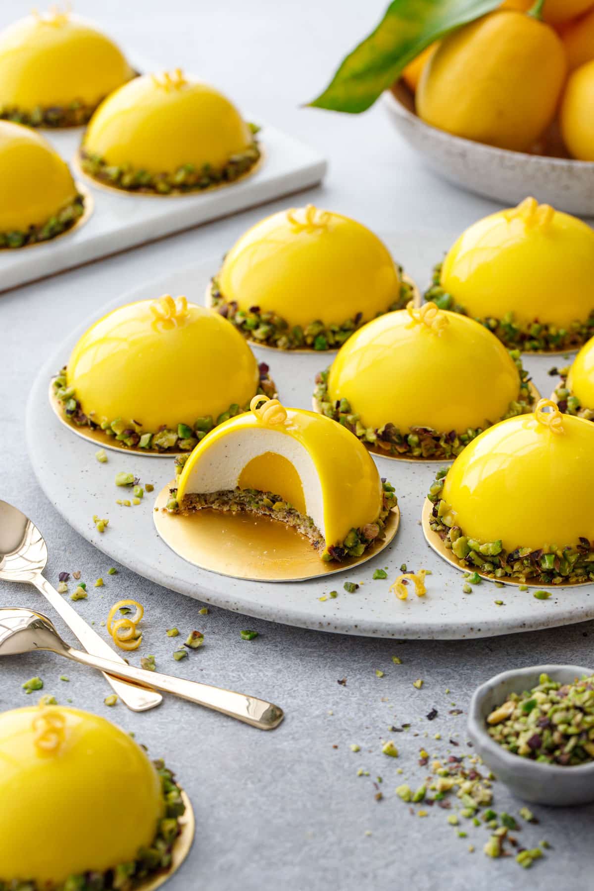 Round plate with bright yellow dome-shaped mousse cakes, one cut to show the layers inside; gold spoons and bowl of lemons in the background.