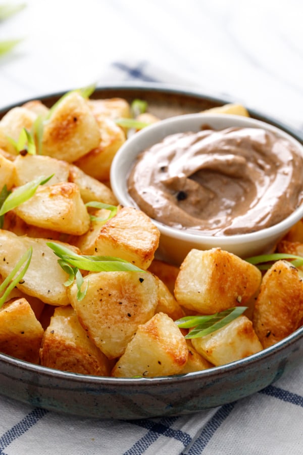 Shallow bowl filled with golden brown crispy potatoes, with a dish of black garlic aioli