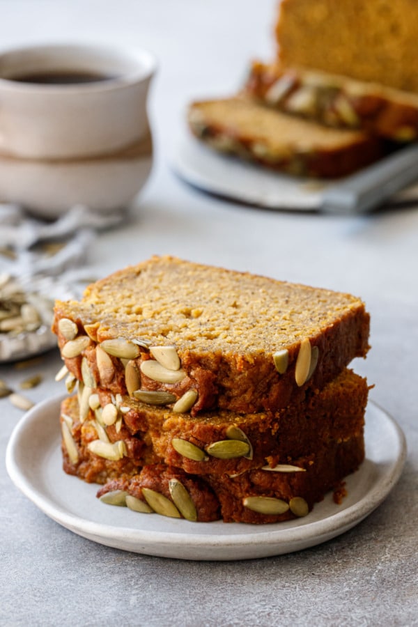 Stack of three slices of Spiced Pumpkin Banana Bread on a ceramic plate, cup of coffee and rest of loaf in the background.