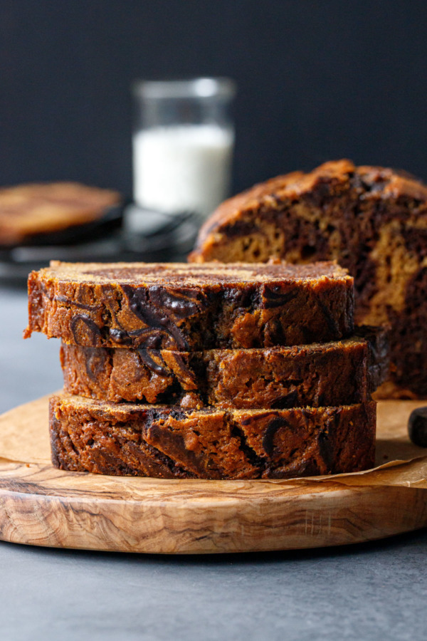 Stack of three slices of Pumpkin Chocolate Swirl Bread on a wooden board, plate and glass of milk in the background