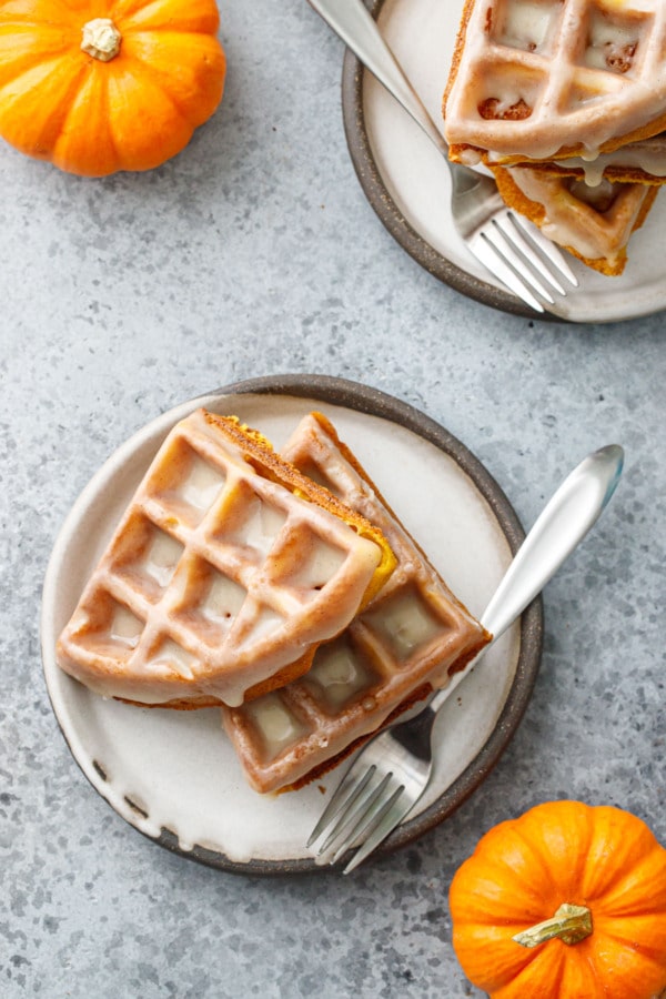 Overhead, two plates of stacked pumpkin waffles with forks, and two mini pumpkins.