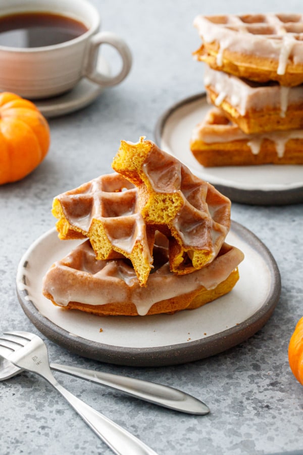 Two plates stacked high with Pumpkin Donut Waffles with Vanilla Cardamom Glaze, one waffle cut in half to show the light and airy texture.