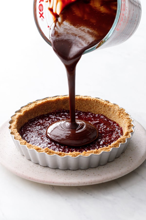 Pouring warm chocolate ganache into a tart crust, with a layer of raspberry jam in the bottom