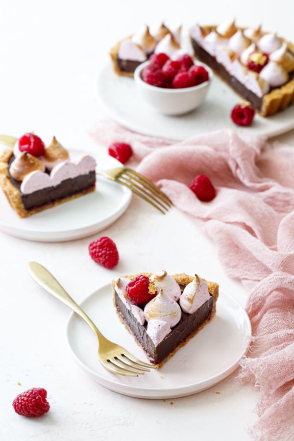 Small pink dessert plates with perfect cut slices of Chocolate Raspberry S'mores Tart; full tart on cake plate in the background with fresh raspberries.