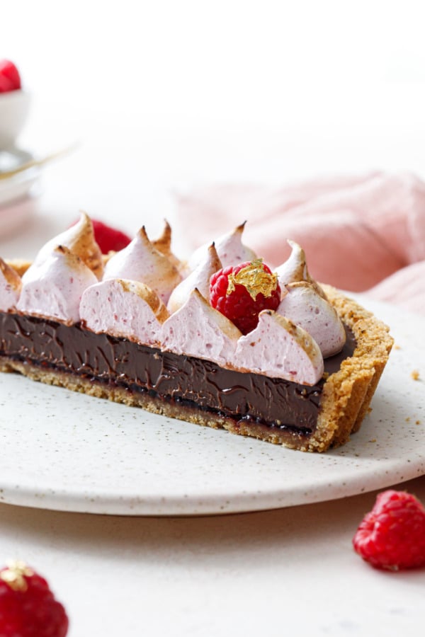 Close up Chocolate Raspberry S'mores Tart cut in half to show the cross-section of layers: graham crust, raspberry jam, chocolate ganache, light pink raspberry meringue.