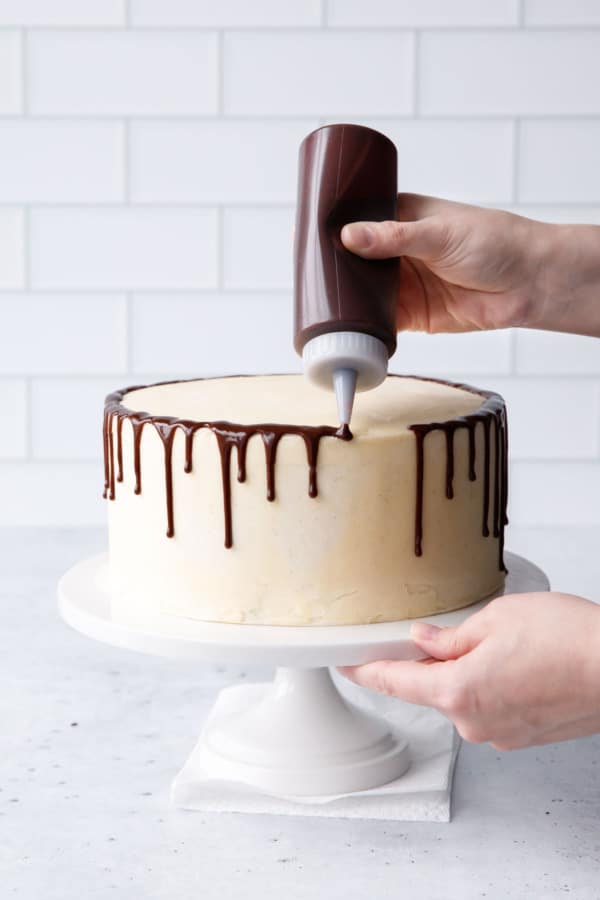 Using a squeeze bottle to drizzle the chocolate glaze around the rim of the frosted cake.