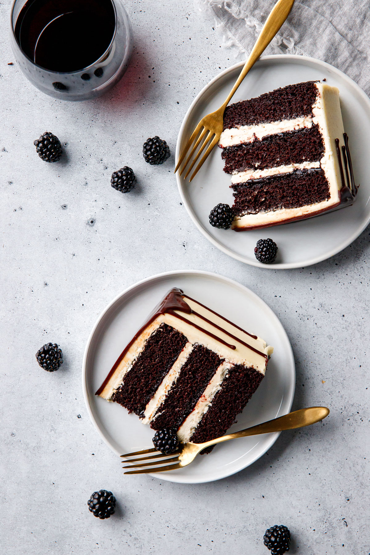 Overhead, two white plates with slices of Blackberry Red Wine Chocolate Cake laying on their sides, with gold forks, glass of red wine, and a few fresh blackberries scattered around.