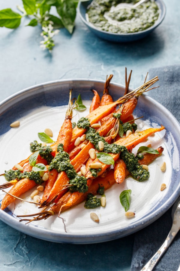 Ceramic platter with pile of roasted carrots, drizzled with pesto and sprinkled with pine nuts.