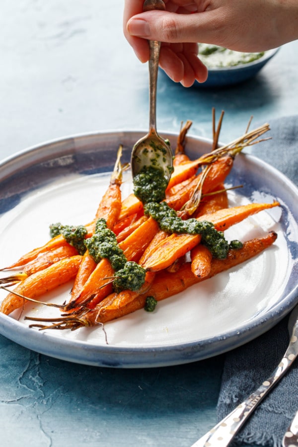 Hand holding spoon drizzling roasted carrots with a basil and carrot top pesto.