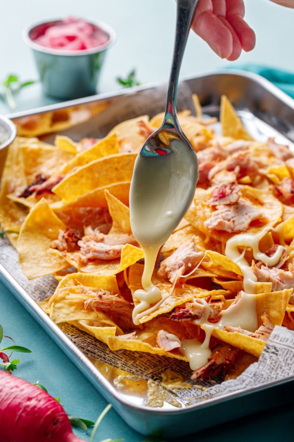Spoon drizzling nacho cheese sauce over a sheet pan of tortilla chips