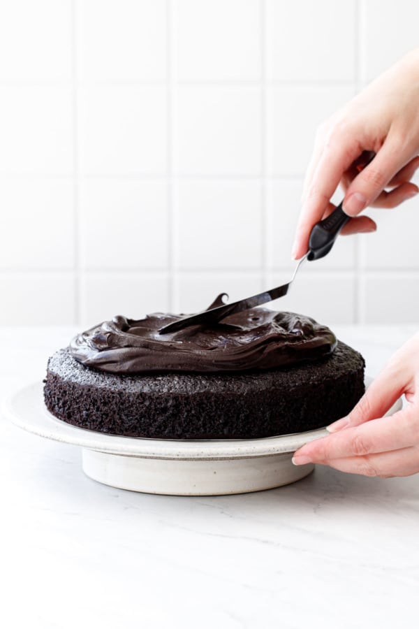 Spreading glossy chocolate frosting onto an extra dark chocolate sour cream cake, on a white cake stand.