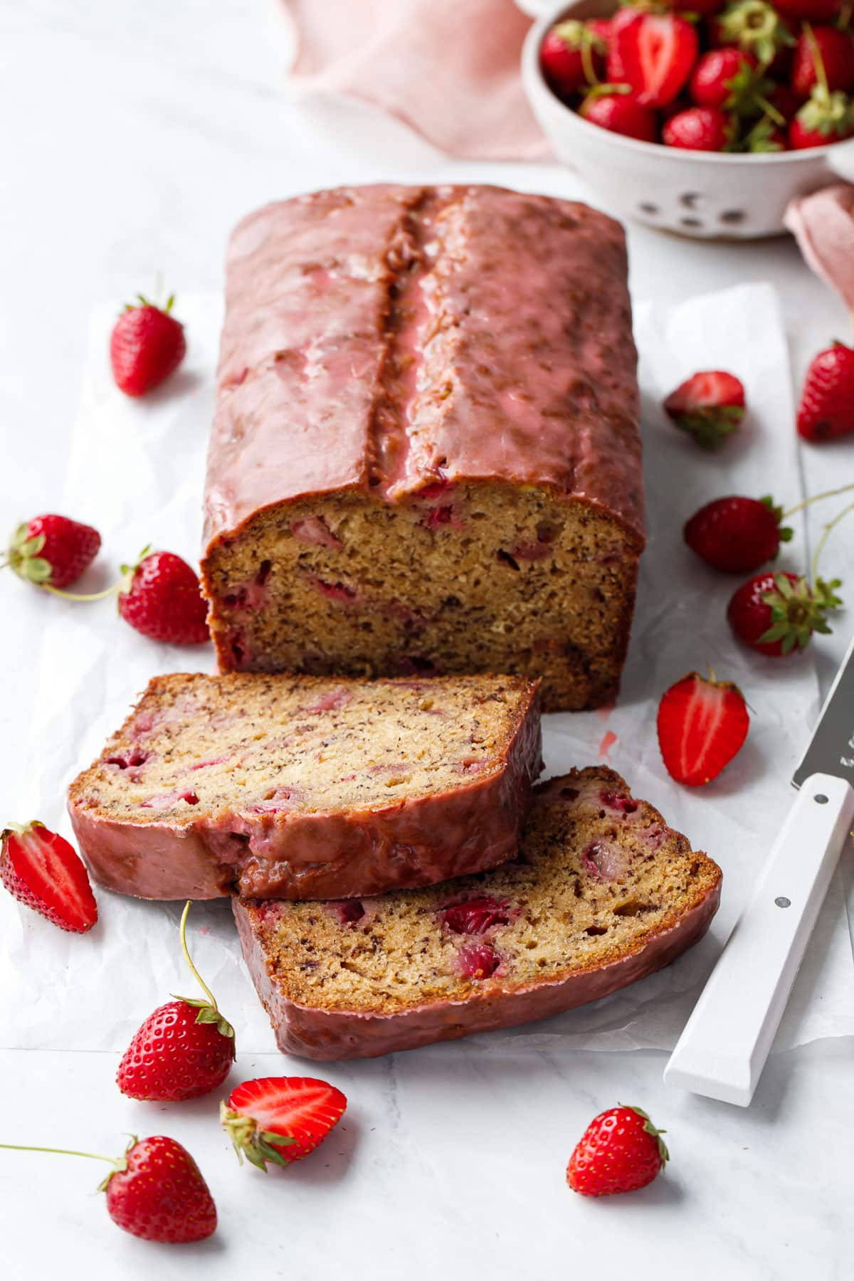 Loaf of Strawberry Banana Bread with a pink glaze, two slices laying down to show the interior texture, and strainer with strawberries, knife, and pink napkin in the background.