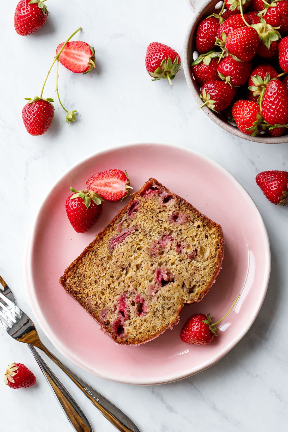 Overhead, slice of Strawberry Banana Bread on a pink plate showing the moist texture and strawberries studded throughout, with fresh strawberries scattered around.