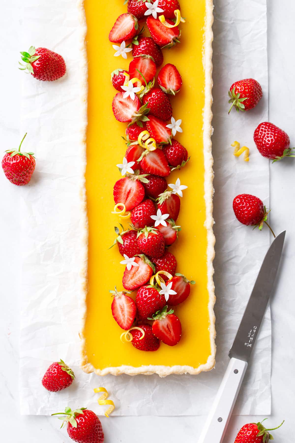 Overhead, rectangular Strawberry Meyer Lemon Tart that is bright yellow with fresh strawberries down the center, with twists of lemon peel and edible flowers.