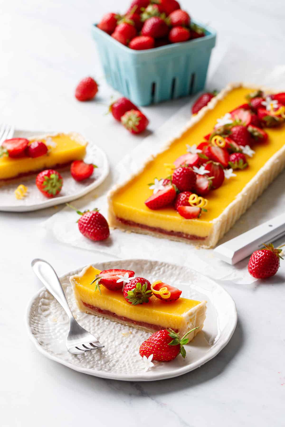 Slice of Strawberry Meyer Lemon Tart on a white plate, garnished with fresh strawberries and white edible flowers.