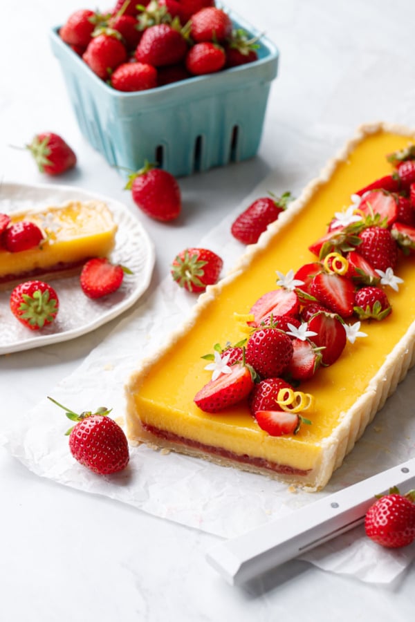 Strawberry Meyer Lemon Tart, cut to show the layers of bright yellow curd and strawberry jam; basket of strawberries and plate in the background.