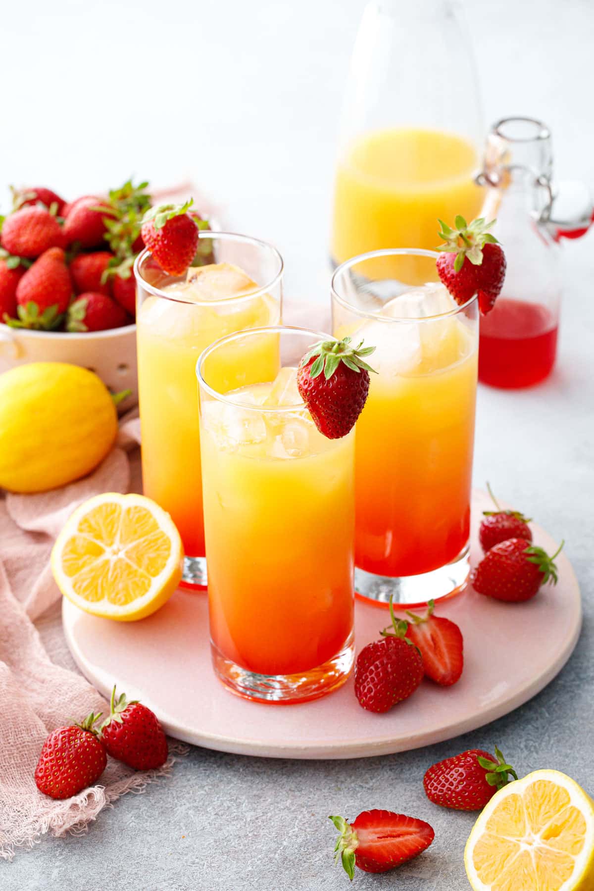Three glasses with Strawberry Passionfruit Lemonade, ombre colored with red at the bottom and yellow at the top, with lemons and strawberries scattered around.